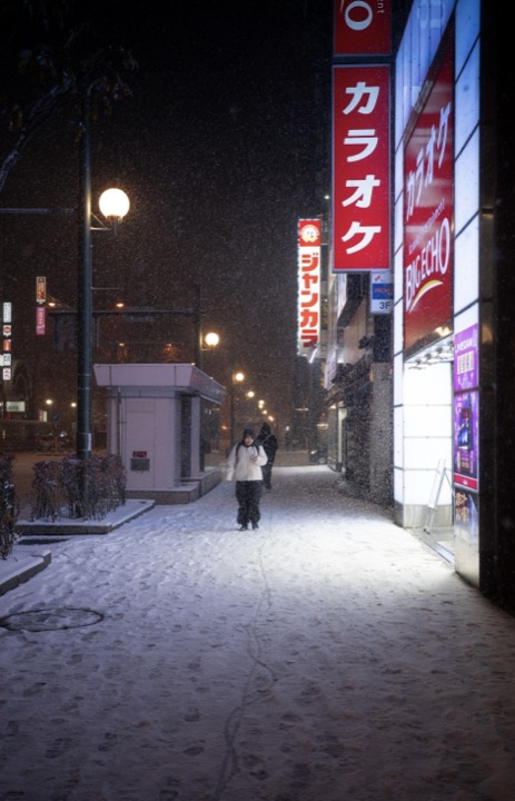 Night-time street in falling snow, a person walking past glowing karaoke signs